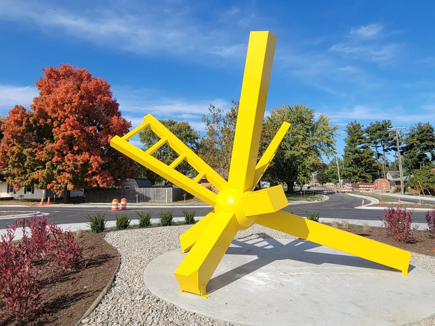 A bright yellow steel sculpture on a landscaped roundabout in Greenwood, with a red maple behind it